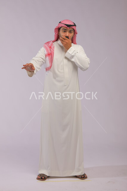 الصدمة بسماع الأخبار والمفاجأة برؤية شيء ما، بورتريه لرجل عربي سعودي خليجي يرتدي الزي التقليدي والشماغ يضع يده على فمه، لغة جسد وإيماءات وجه و يدين تدل على الدهشة و الذهول، صورة الجسم بالطول الكامل، خلفية رمادية.