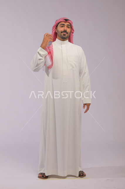 عطورات خليجية سعودية, مفهوم الاناقة والاهتمام بالمظهر الخارجي, استخدام الزيوت العطرية الطبيعية, بورتريه لرجل عربي خليجي سعودي يرتدي الثوب والشماغ يقوم بوضع العطر, التسويق لمنتجات العطور, صورة الجسم بالكامل, خلفية رمادية