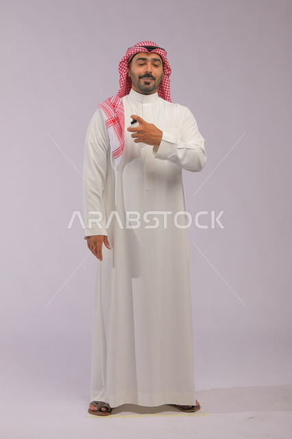 عطورات خليجية سعودية, مفهوم الاناقة والاهتمام بالمظهر الخارجي, استخدام الزيوت العطرية الطبيعية, بورتريه لرجل عربي خليجي سعودي يرتدي الثوب والشماغ يقوم بوضع العطر, التسويق لمنتجات العطور, صورة الجسم بالكامل, خلفية رمادية