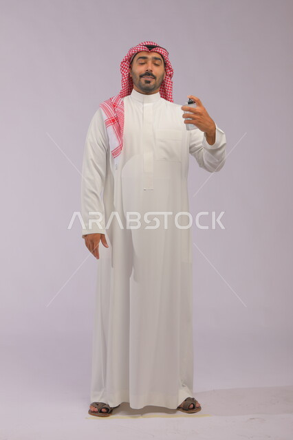 التسويق لمنتجات العطور, عطورات خليجية سعودية, مفهوم الاناقة والاهتمام بالمظهر الخارجي, استخدام الزيوت العطرية الطبيعية, بورتريه لرجل عربي خليجي سعودي يرتدي الثوب والشماغ يقوم بوضع العطر, صورة الجسم بالكامل, خلفية رمادية