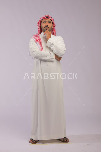 إيماءات التمعن والتركيز، التأمل وشرود الذهن والتخطيط للمستقبل, بورتريه لرجل عربي خليجي سعودي يرتدي الزي التقليدي يقوم بالتفكير لإيجاد حل لمشكلة ما, صورة الجسم بالطول الكامل، خلفية رمادية .