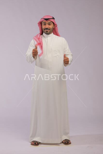 علامة الجودة والامتيازوالتميز، بورتريه لرجل عربي خليجي سعودي يرتدي الثوب التقليدي والشماغ ويشير بيده بإيماءات وتعابير وجه و يدين تدل على الاعجاب، التعبير عن الجودة والاتقان، القبول والموافقة والرضا، صورة الجسم بالطول الكامل، خلفية رمادية.