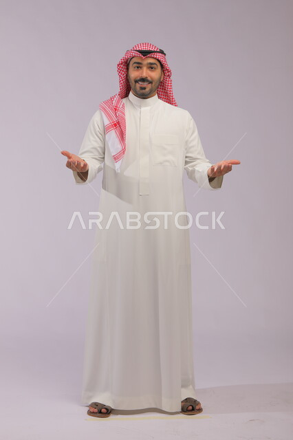 تعابير الاعجاب والمدح لشيء جميل، بورتريه لرجل عربي خليجي سعودي مبتسم يرتدي الشماغ والثوب التقليدي يقوم بفتح كفيه والنظر إلى الكاميرا بإيماءات السرور والبشاشة والترحيب، صورة الجسم بالطول الكامل، خلفية رمادية.