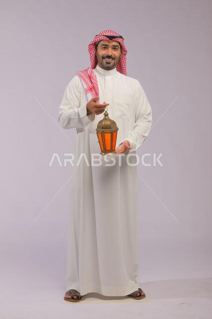 الاستعداد لقدوم شهر رمضان, المناسبات الدينية والأعياد الإسلامية, بورتريه لشاب عربي خليجي سعودي مبتسم يرتدي الثوب التقليدي والشماغ يحمل الفانوس المضيء, تحف وزينة الاحتفالات في السعودية, صورة الجسم بالكامل, اسلاميات وعبادة, خلفية رمادية