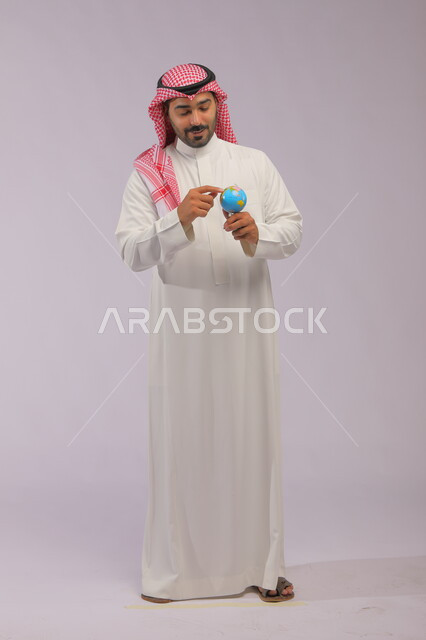 خريطة العالم الكروية, مفهوم البحث والاستكشاف حول العالم, بورتريه لرجل عربي خليجي سعودي مبتسم يرتدي الثوب التقليدي والشماغ يحمل مجسم الكرة الارضية, الاشارة إلى المجسم بإيماءات الاهتمام والتركيز, صورة الجسم بالطول الكامل, خلفية رمادية