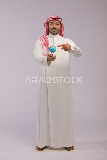 The concept of research and exploration around the world, a portrait of a smiling Saudi Gulf Arab man wearing a traditional thobe and shemagh holding a globe, pointing to the globe and looking at the camera, spherical world map, full-length body portrait, gray background