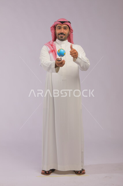 The concept of research and exploration around the world, portrait of a smiling Saudi Gulf Arab man wearing a traditional thobe and shemagh holding a globe, looking at the camera with quality gestures, spherical world map, full-length body portrait, gray background
