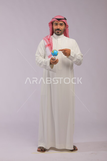 A spherical world map, the concept of research and exploration around the world, a portrait of a smiling Saudi Arabian Gulf man wearing a traditional thobe and shemagh holding a globe, pointing to the globe and looking at the camera, full-length body portrait, gray background