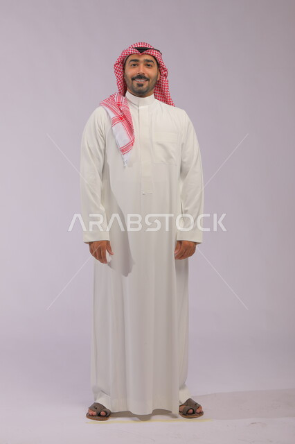 مفهوم الاهتمام بالمظهر الخارجي و الرجولة والأناقه، ايماءات البشاشة والسرور، بورتريه لرجل عربي خليجي سعودي مبتسم يرتدي الشماغ والثوب التقليدي يقف باستقامة وثقة وينظر إلى الكاميرا، صورة الجسم بالطول الكامل، خلفية رمادية.