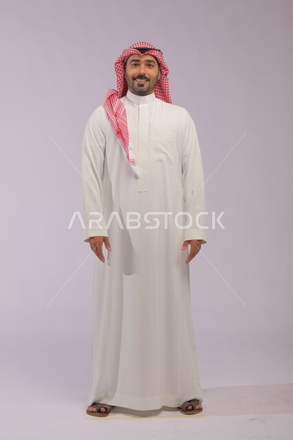 مفهوم الاهتمام بالمظهر الخارجي و الرجولة والأناقه، ايماءات البشاشة والسرور، بورتريه لرجل عربي خليجي سعودي مبتسم يرتدي الشماغ والثوب التقليدي يقف باستقامة وثقة وينظر إلى الكاميرا، صورة الجسم بالطول الكامل، خلفية رمادية.