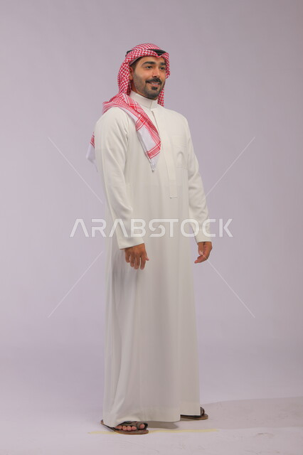 مفهوم الاهتمام بالمظهر الخارجي و الرجولة والأناقه، ايماءات البشاشة والسرور، بورتريه لرجل عربي خليجي سعودي مبتسم يرتدي الشماغ والثوب التقليدي يقف باستقامة وثقة، صورة الجسم بالطول الكامل، خلفية رمادية.