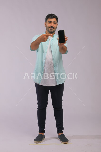 Electronic marketing using the Internet, a portrait of a Saudi Gulf Arab man wearing casual summer clothes, pointing with his hand to a blank black advertising screen on the mobile phone, integrating technology and technology into practical life, full-length body image, white background.