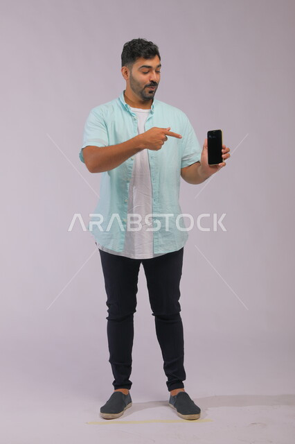 استعمال التقنيات الحديثة في التسويق الالكتروني، بورتريه لرجل عربي سعودي خليجي يرتدي ملابس كاجوال صيفية يشير بيده الى شاشة اعلانية سوداء فارغة على الجوال، دور التقنية الحديثة في توفير راحة أكبر للأفراد، صورة الجسم بالطول الكامل، خلفية بيضاء.