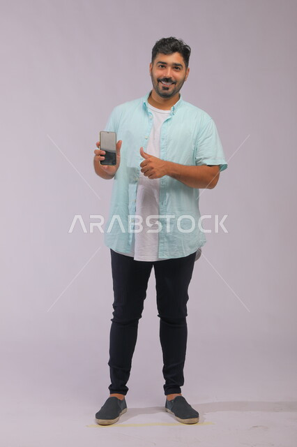 ايماءات الاعجاب باستخدام التقنية الحديثة، بورتريه لرجل عربي سعودي خليجي يرتدي ملابس كاجوال صيفية يمسك بيده الجوال بشاشة اعلانية سوداء فارغة، مفهوم التسويق الالكتروني،  صورة الجسم بالطول الكامل، خلفية بيضاء.