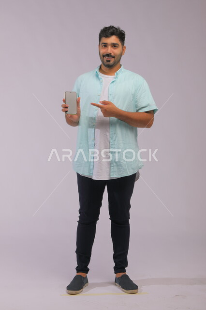 The concept of electronic marketing, a portrait of a Saudi Gulf Arab man wearing casual summer clothes, pointing with his hand to a blank white advertising screen on a mobile phone, the role of modern technology in providing greater comfort for individuals, full-length body image, white background.