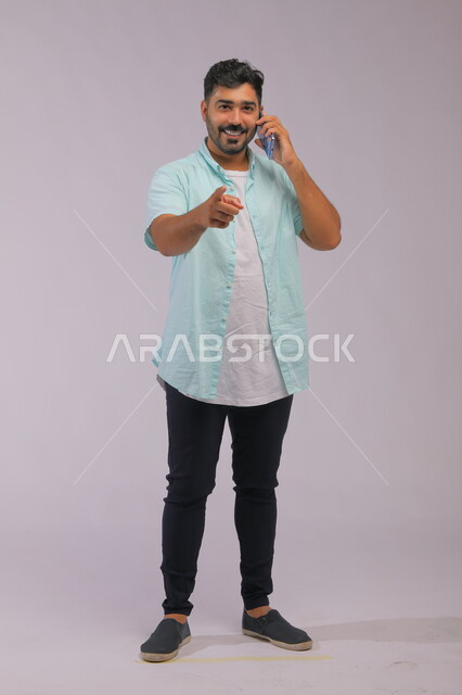Making a business call, the role of modern technology in providing greater comfort for individuals, improving the quality of life using the mobile phone, a portrait of a Saudi Gulf Arab man wearing casual summer clothes, talking using the mobile phone and pointing with his hand forward, full-length body portrait, white background.