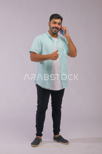 ايماءات الموافقة على الكلام، إجراء مكالمة عمل، دور التقنية الحديثة في توفير راحة أكبر للأفراد، تحسين جودة الحياة باستخدام الجوال، بورتريه لرجل عربي سعودي خليجي يرتدي ملابس كاجوال صيفية ويتحدث باستخدام الهاتف المحمول، صورة الجسم بالطول الكامل، خلفية بيضاء.