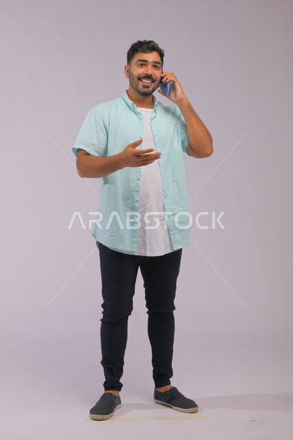 إجراء مكالمة عمل، دور التقنية الحديثة في توفير راحة أكبر للأفراد، ايماءات التفاعل اثناء المكالمة الهاتفية، بورتريه لرجل عربي سعودي خليجي يرتدي ملابس كاجوال صيفية ويتحدث باستخدام الهاتف المحمول، صورة الجسم بالطول الكامل، خلفية بيضاء.