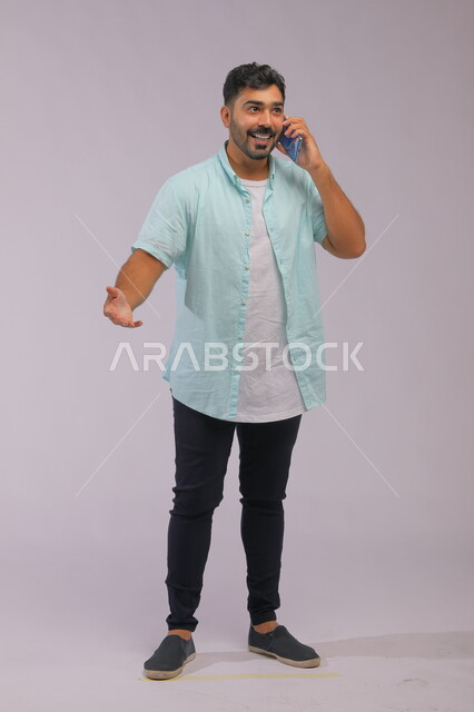 Communicating with family and friends easily, interaction gestures during a phone call, portrait of a Saudi Gulf Arab man wearing casual summer clothes and talking using a mobile phone, full-length body portrait, white background.