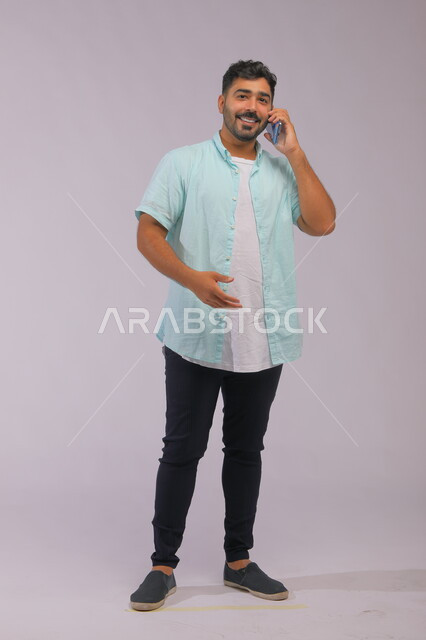 ايماءات التفاعل اثناء المكالمة الهاتفية، التواصل مع العائلة والأصدقاء بسهولة، بورتريه لرجل عربي سعودي خليجي يرتدي ملابس كاجوال صيفية ويتحدث باستخدام الهاتف المحمول، صورة الجسم بالطول الكامل، خلفية بيضاء.