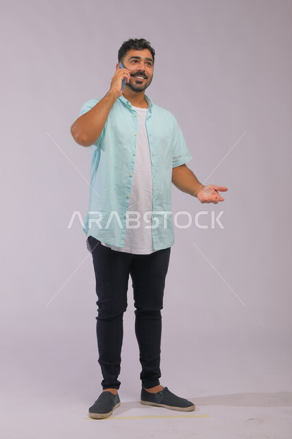 ايماءات التفاعل اثناء المكالمة الهاتفية، التواصل مع العائلة والأصدقاء بسهولة، بورتريه لرجل عربي سعودي خليجي يرتدي ملابس كاجوال صيفية ويتحدث باستخدام الهاتف المحمول، صورة الجسم بالطول الكامل، خلفية بيضاء.