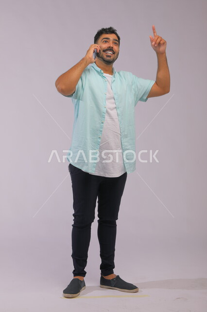 التحدث مع الأصدقاء عبر الجوال، التواصل مع العالم بسهولة، بورتريه لشاب عربي سعودي خليجي يرتدي الزي الكاجول ويقوم باجراء مكالمة هاتفية، إستخدام تطبيقات التواصل الاجتماعي، صورة الجسم بالكامل، خلفية بيضاء.