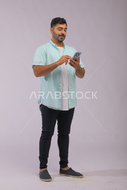 التسوق بسهولة وراحة عبر الانترنت، التواصل مع العالم بسهولة، بورتريه لشاب عربي سعودي خليجي يرتدي الزي الكاجول ويمسك بيده الهاتف المحمول، إستخدام برامج الهاتف الذكي، صورة الجسم بالكامل، خلفية بيضاء.