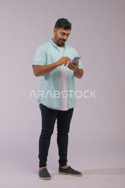 التواصل مع الأصدقاء عبر مواقع التواصل الاجتماعي، متابعة الاحداث العالمية باستخدام الهاتف المحمول، بورتريه لشاب عربي سعودي خليجي يرتدي الزي الكاجول ويمسك بيده الجوال، صورة الجسم بالكامل، خلفية بيضاء