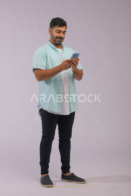 التواصل مع الأصدقاء عبر مواقع التواصل الاجتماعي، متابعة الاحداث العالمية باستخدام الهاتف المحمول، بورتريه لشاب عربي سعودي خليجي يرتدي الزي الكاجول ويمسك بيدها الجوال ، صورة الجسم بالكامل، خلفية بيضاء