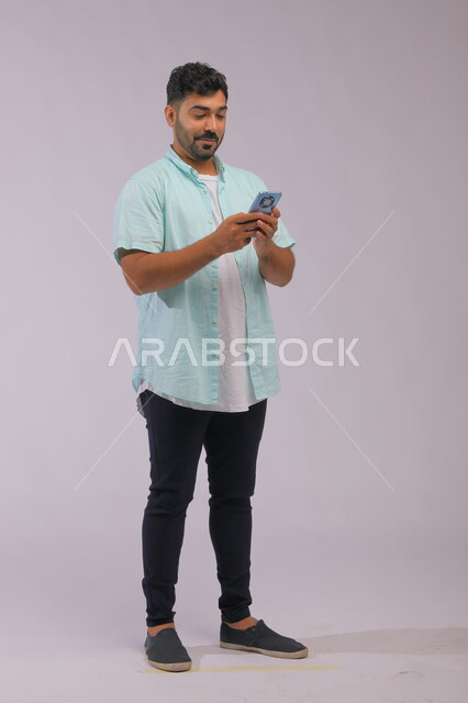 متابعة الاحداث العالمية باستخدام الجوال، بورتريه لشاب عربي سعودي خليجي يرتدي الزي الكاجول ويمسك بيده الهاتف المحمول، التواصل مع الأصدقاء عبر مواقع التواصل الاجتماعي، صورة الجسم بالكامل، خلفية بيضاء
