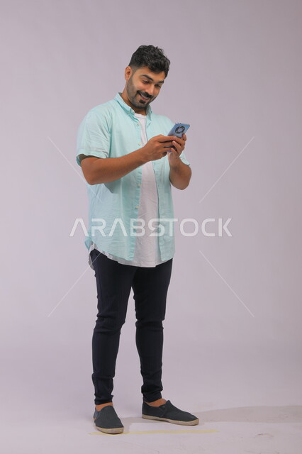متابعة الاحداث العالمية باستخدام الجوال، بورتريه لشاب عربي سعودي خليجي يرتدي الزي الكاجول ويمسك بيده الهاتف المحمول، التواصل مع الأصدقاء عبر مواقع التواصل الاجتماعي، صورة الجسم بالكامل، خلفية بيضاء.