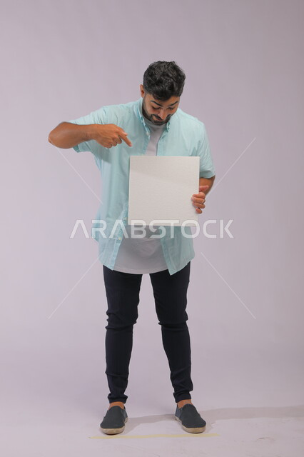 إعلانات وعروض تسويقية، استخدام لوحات توضيحية ووسائل تعليمية، موك اب مربع أبيض، بورتريه لشاب عربي خليجي سعودي مبتسم يرتدي الزي الكاجوال و يشير بيده إلى لوحه بيضاء مربعه فارغة بإيماءات التعجب والذهول، صورة الجسم بالكامل، خلفية رماديه.