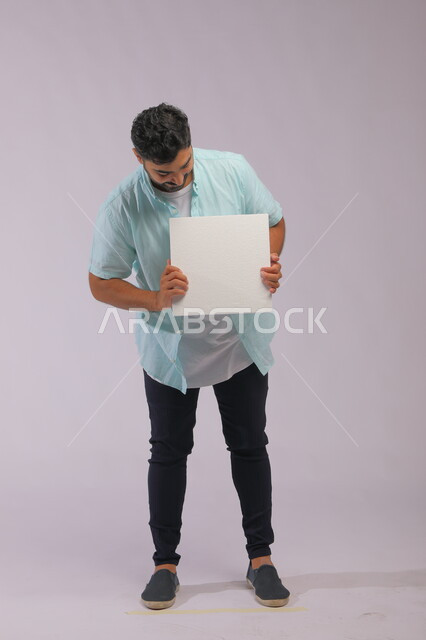 موك اب مربع أبيض، إعلانات وعروض تسويقية، استخدام لوحات توضيحية ووسائل تعليمية، بورتريه لشاب عربي خليجي سعودي مبتسم يرتدي الزي الكاجوال ويمسك بيديه لوحه بيضاء مربعه فارغة، صورة الجسم بالكامل، خلفية رماديه.