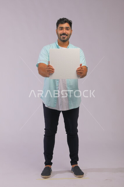 النظر إلى الكاميرا بإيماءات وجه تدل على السرور، موك اب مربع أبيض، إعلانات وعروض تسويقية، استخدام لوحات توضيحية ووسائل تعليمية، بورتريه لشاب عربي خليجي سعودي مبتسم يرتدي الزي الكاجوال ويعرض لوحه بيضاء مربعه فارغة، صورة الجسم بالكامل، خلفية رماديه.