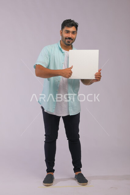 النظر إلى الكاميرا بلغة جسد وإشارات تدل على الإعجاب والقبول، موك اب مربع أبيض، إعلانات وعروض تسويقية، استخدام لوحات توضيحية ووسائل تعليمية، بورتريه لشاب عربي خليجي سعودي مبتسم يرتدي الزي الكاجوال ويعرض لوحه بيضاء مربعه فارغة، صورة الجسم بالكامل، خلفية رماديه.
