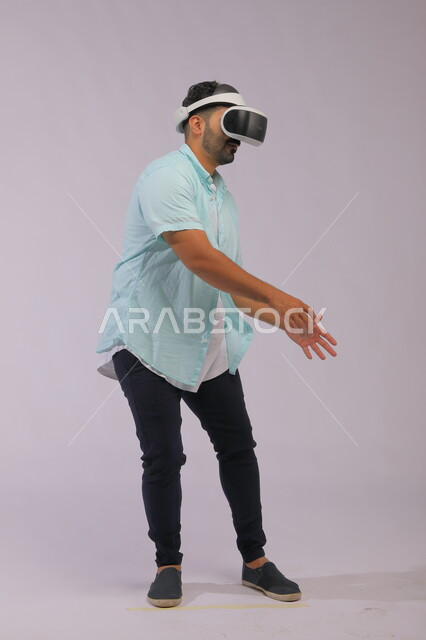 استخدام التكنولوجيا والتقنية الحديثة، بورتريه لرجل عربي سعودي خليجي يرتدي ملابس كاجوال صيفية ويضع نظارات الواقع الافتراضي، ايماءات التفاعل مع الشخصيات في اللعبة، صورة الجسم بالطوال الكامل، خلفية بيضاء
