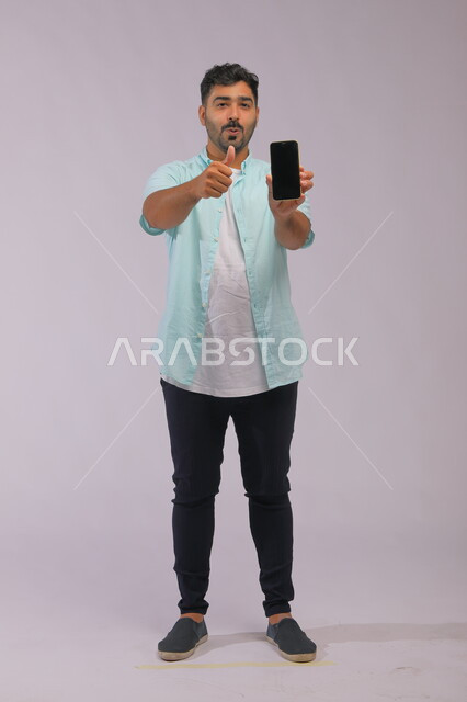 ايماءات الاعجاب باستخدام التقنية الحديثة، بورتريه لرجل عربي سعودي خليجي يرتدي ملابس كاجوال صيفية يمسك بيده الجوال بشاشة اعلانية سوداء فارغة، مفهوم التسويق الالكتروني،  صورة الجسم بالطول الكامل، خلفية بيضاء.