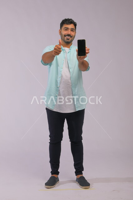 Gestures of admiration using modern technology, a portrait of a Saudi Gulf Arab man wearing casual summer clothes, holding a mobile phone in his hand with a blank black advertising screen, electronic marketing concept, full-length body image, white background.