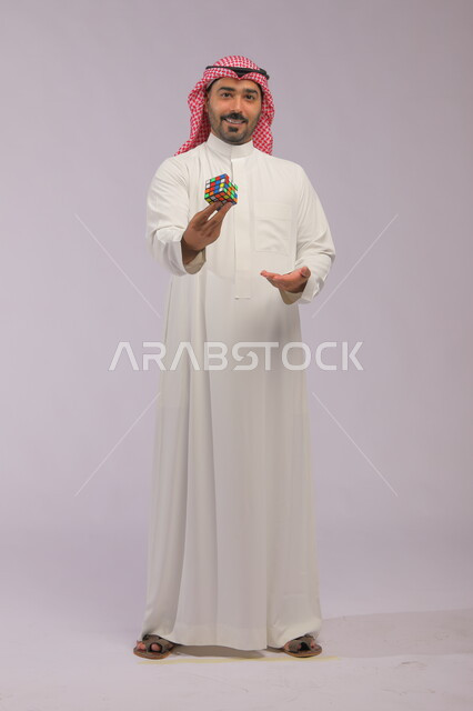 ألعاب تنمية الذكاء والمهارات العقليه, ممارسة الألعاب التعليمية الترفيهية, عرض تجربة اللعب بالروبيك كيوب, بورتريه لشاب عربي خليجي سعودي يرتدي الثوب التقليدي والشماغ يمسك بيده مكعب مغناطيس السرعة وينظر إلى الكاميرا, صورة الجسم بالطول الكامل, خلفية رمادية