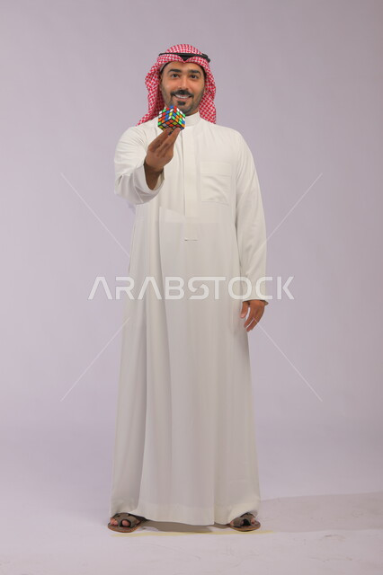 عرض تجربة اللعب بالروبيك كيوب, ممارسة الألعاب التعليمية الترفيهية, بورتريه لشاب عربي خليجي سعودي يرتدي الثوب التقليدي والشماغ يمسك بيده مكعب مغناطيس السرعة وينظر إلى الكاميرا, ألعاب تنمية الذكاء والمهارات العقليه, صورة الجسم بالطول الكامل, خلفية رمادية