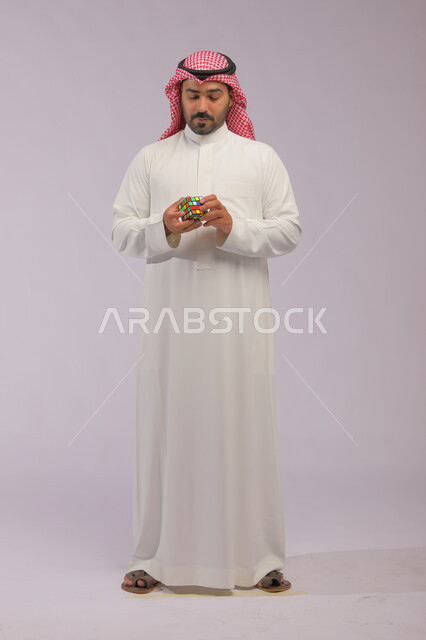 ألعاب تنمية الذكاء والمهارات العقليه, ممارسة الألعاب التعليمية الترفيهية, عرض تجربة اللعب بالروبيك كيوب, بورتريه لشاب عربي خليجي سعودي يرتدي الثوب التقليدي والشماغ يمسك بيده مكعب مغناطيس السرعة, صورة الجسم بالطول الكامل, خلفية رمادية