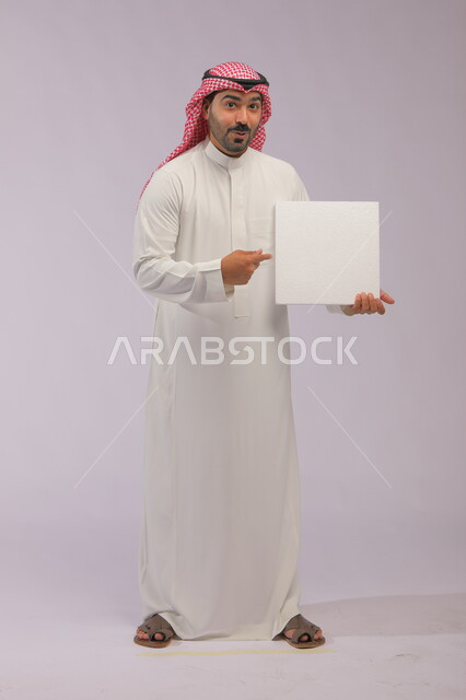 إعلانات وعروض تسويقية، استخدام لوحات توضيحية ووسائل تعليمية، موك اب مربع أبيض، بورتريه لشاب عربي خليجي سعودي يرتدي الثوب التقليدي والشماغ يشير بيده إلى لوحه بيضاء مربعه فارغة بإيماءات الدهشة والذهول، صورة الجسم بالكامل، خلفية رماديه