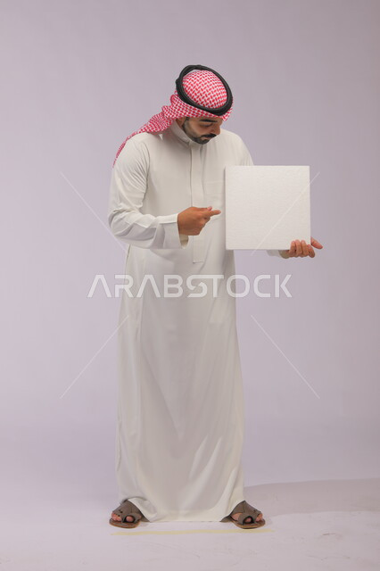 عروض وإعلانات تسويقية, موك اب مربع أبيض، استخدام لوحات توضيحية ووسائل تعليمية، بورتريه لشاب عربي خليجي سعودي مبتسم يرتدي الثوب التقليدي والشماغ يمسك بيديه لوحه بيضاء مربعه فارغة، صورة الجسم بالطول الكامل، خلفية رماديه