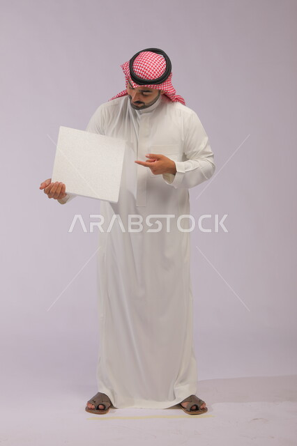 موك اب مربع أبيض، إعلانات وعروض تسويقية، استخدام لوحات توضيحية ووسائل تعليمية، بورتريه لشاب عربي خليجي سعودي مبتسم يرتدي الثوب التقليدي والشماغ يمسك بيديه لوحه بيضاء مربعه فارغة، صورة الجسم بالطول الكامل، خلفية رماديه