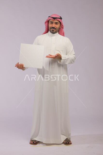 استخدام لوحات توضيحية ووسائل تعليمية، إعلانات وعروض تسويقية، موك اب مربع أبيض، بورتريه لشاب عربي خليجي سعودي مبتسم يرتدي الثوب التقليدي والشماغ يشير بيده إلى لوحه بيضاء مربعه فارغة وينظر إلى الكاميرا، صورة الجسم بالكامل، خلفية رماديه