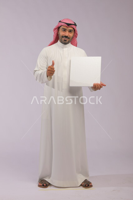 Looking at the camera with gestures of acceptance and goodness, a white square mockup, advertisements and marketing presentations, use of explanatory panels and teaching aids, a portrait of a smiling Gulf Arab Saudi man wearing a traditional thobe and shemagh displaying a blank white board, full-length body portrait, gray background.