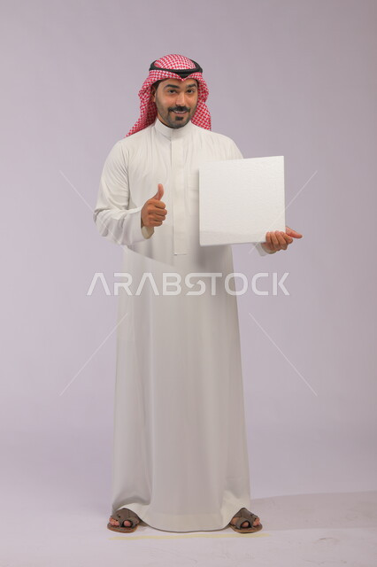 النظر إلى الكاميرا بإيماءات القبول والجودة، موك اب مربع أبيض، إعلانات وعروض تسويقية، استخدام لوحات توضيحية ووسائل تعليمية، بورتريه لشاب عربي خليجي سعودي مبتسم يرتدي الثوب التقليدي والشماغ يعرض لوحه بيضاء فارغة، صورة الجسم بالطول الكامل، خلفية رماديه