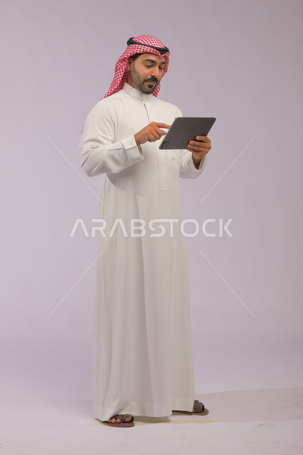 البحث عن فرص عمل عبر الانترنت, التواصل مع الاصدقاء وتصفح مواقع التواصل الاجتماعي, بورتريه لرجل عربي خليجي سعودي يرتدي الثوب التقليدي والشماغ يمسك جهاز التابليت, صورة الجسم بالطول الكامل, خلفية رمادية