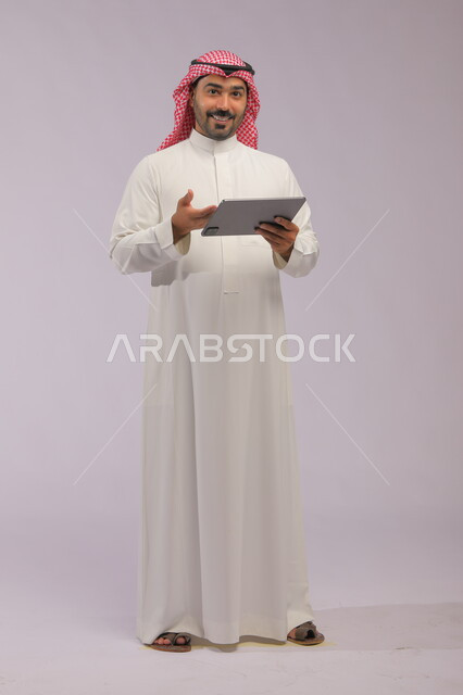 التسويق الالكتروني الذكي والآمن عبر الانترنت, استخدام تطبيقات وتقنيات الأجهزة الحديثة, بورتريه لرجل عربي خليجي سعودي مبتسم يرتدي الثوب التقليدي والشماغ يحمل الجهاز اللوحي, الدردشة والتواصل مع الاصدقاء, صورة الجسم بالطول الكامل, خلفية رمادية
