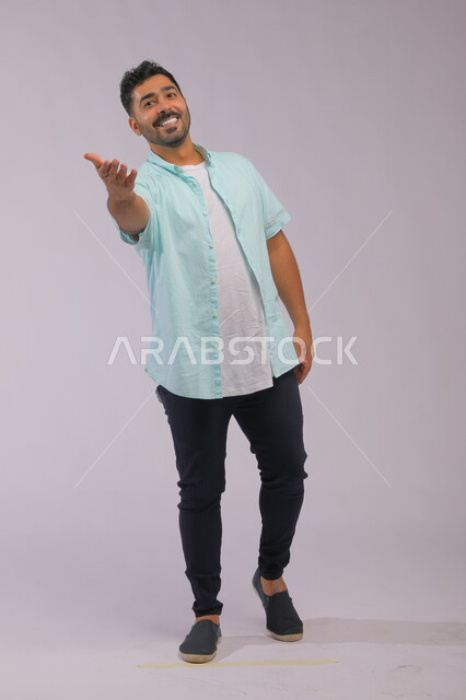 الترحيب والإكرام والاستقبال بحب، إيماءات الفرح والسرور والبشاشة، بورتريه لشاب عربي خليجي سعودي مبتسم يرتدي الزي الكاجوال بلغة جسد تدل على عرض المساعدة للإمساك بيد امرأة، صورة الجسم بالطول الكامل، خلفية رمادية. 
